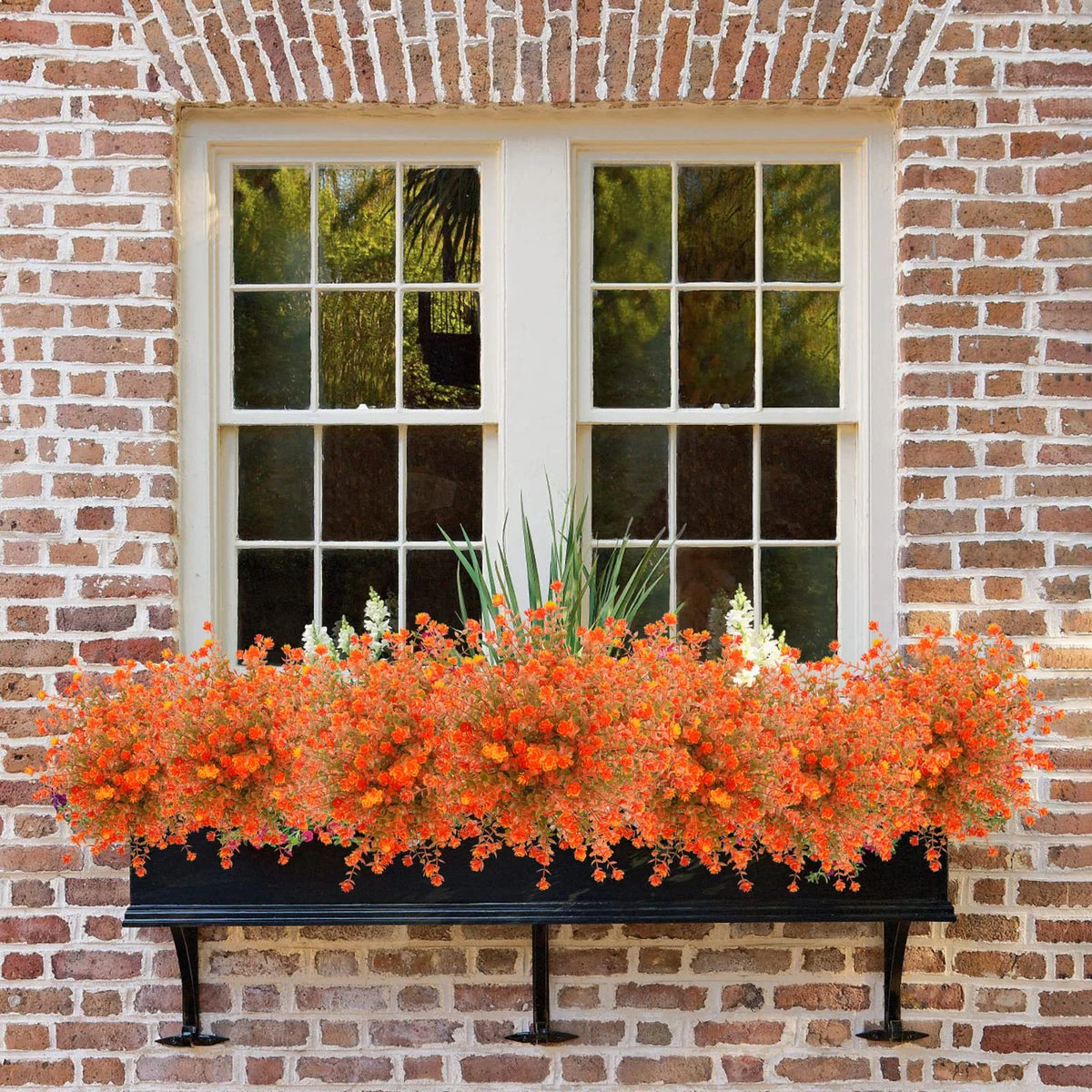 An orange and white artificial daisy bouquet displayed in a black planter on a brick wall windowsill.