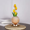 A vase holds three yellow tulips, set on a woven mat and resting on a dark surface.