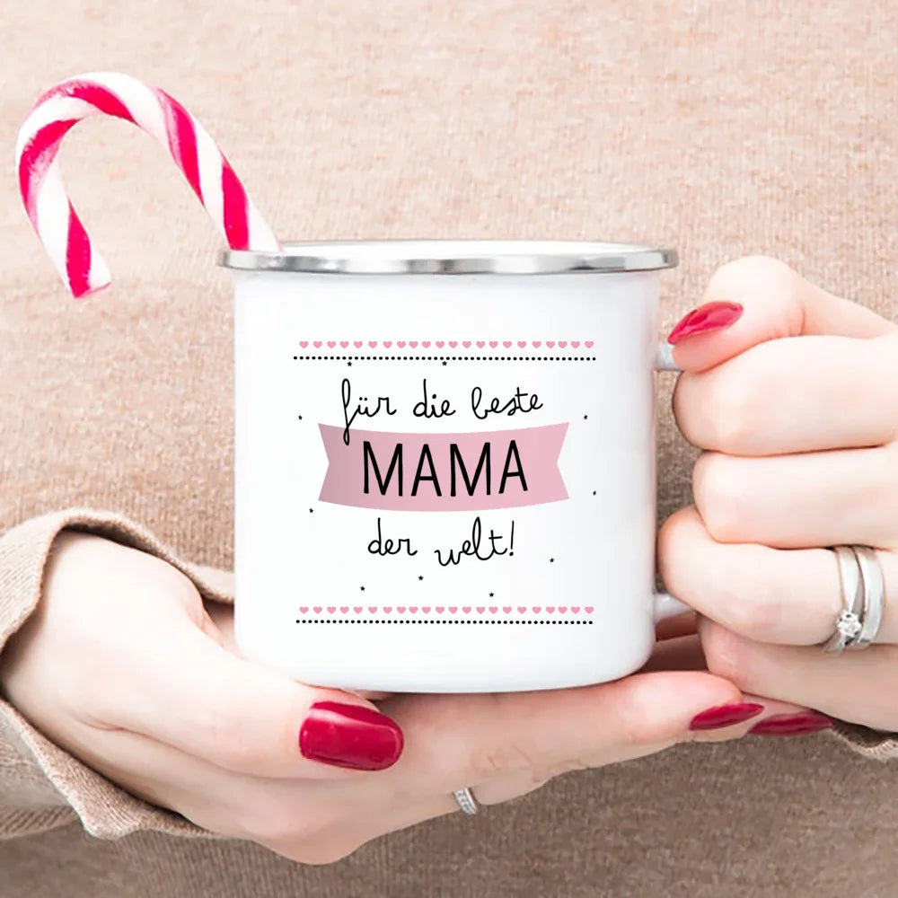 A white enamel mug with pink text that reads für die beste mama der welt, held by a person and with a red and white striped c