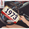 A distressed vintage metal tin sign with the year 1973 is held in a hand, placed on a car steering wheel.