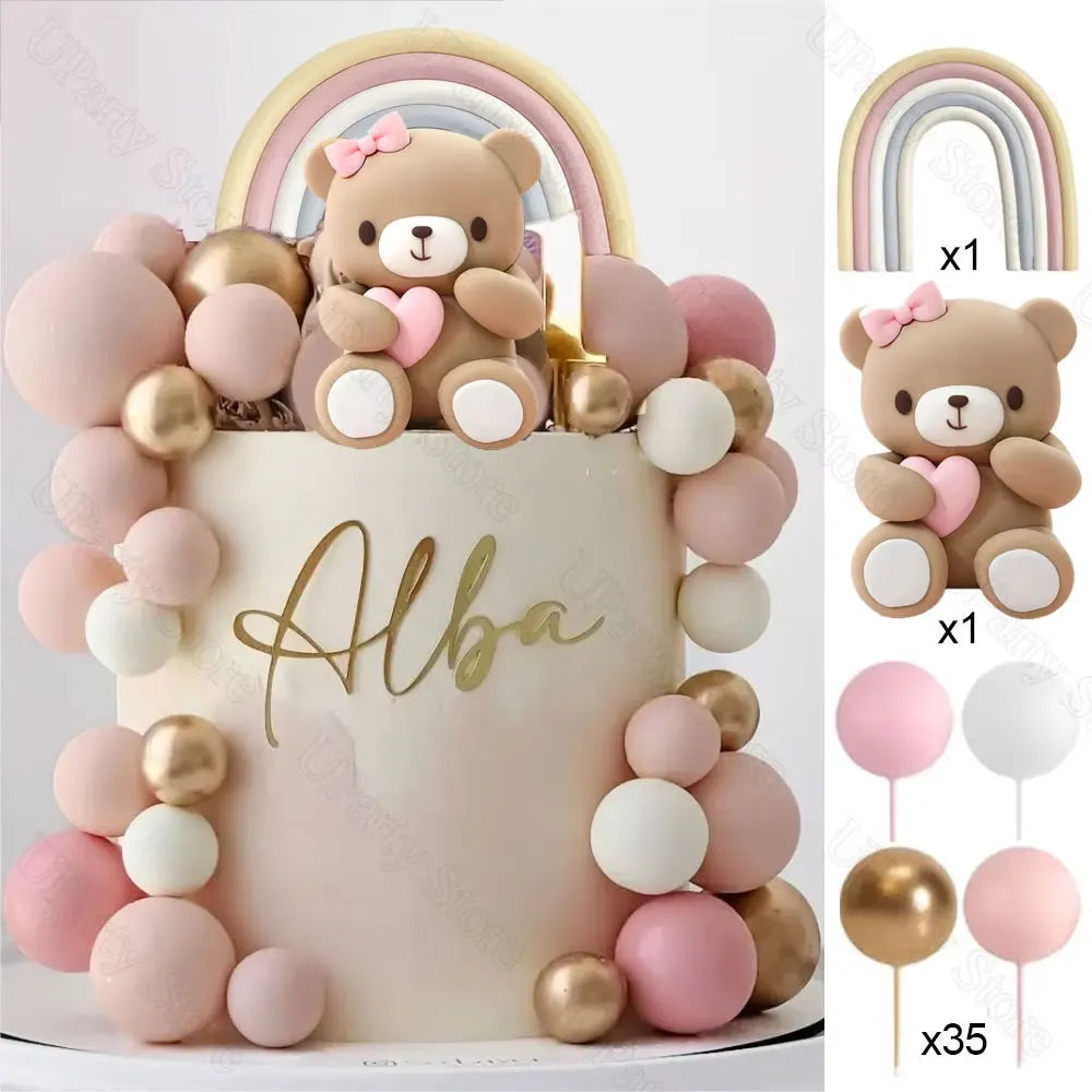 A white cake decorated with pink and gold ball decorations and a teddy bear topper, surrounded by rainbow-colored balloons.