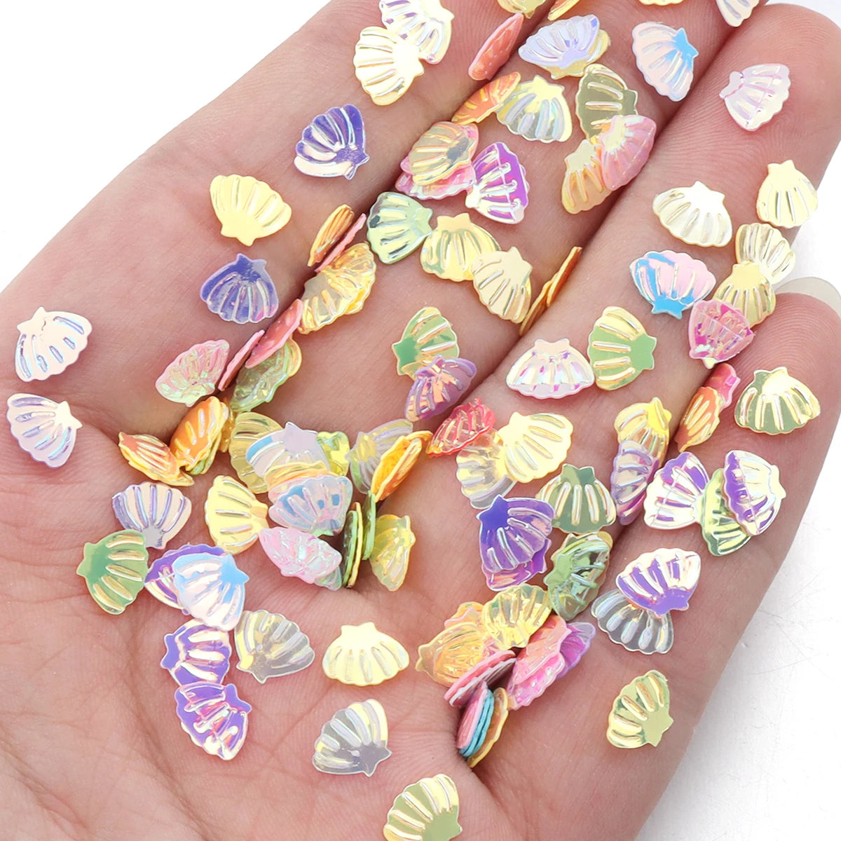 A hand displays various colorful sea shell PVC sequins for nail art and craft decoration.