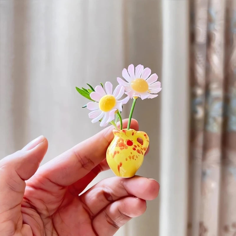 A hand holds a yellow ceramic vase with pink daisy flowers against a light background.