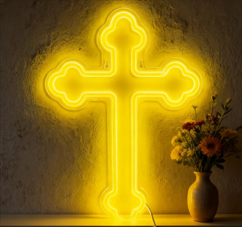 A bright yellow neon cross wall sign illuminates a textured wall, resting on a wooden surface.