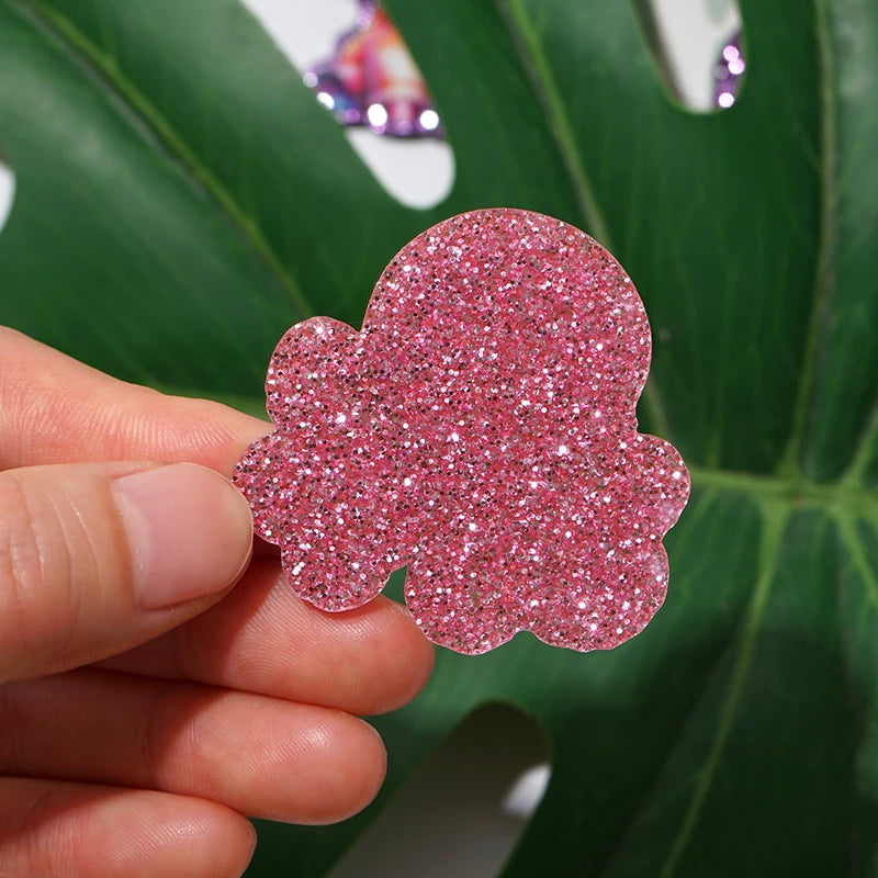 A hand holds a pink glitter acrylic charm shaped like a cloud against a green leaf background.