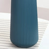 A tall, ribbed, dark teal plastic vase standing on a white surface with a black grid pattern.