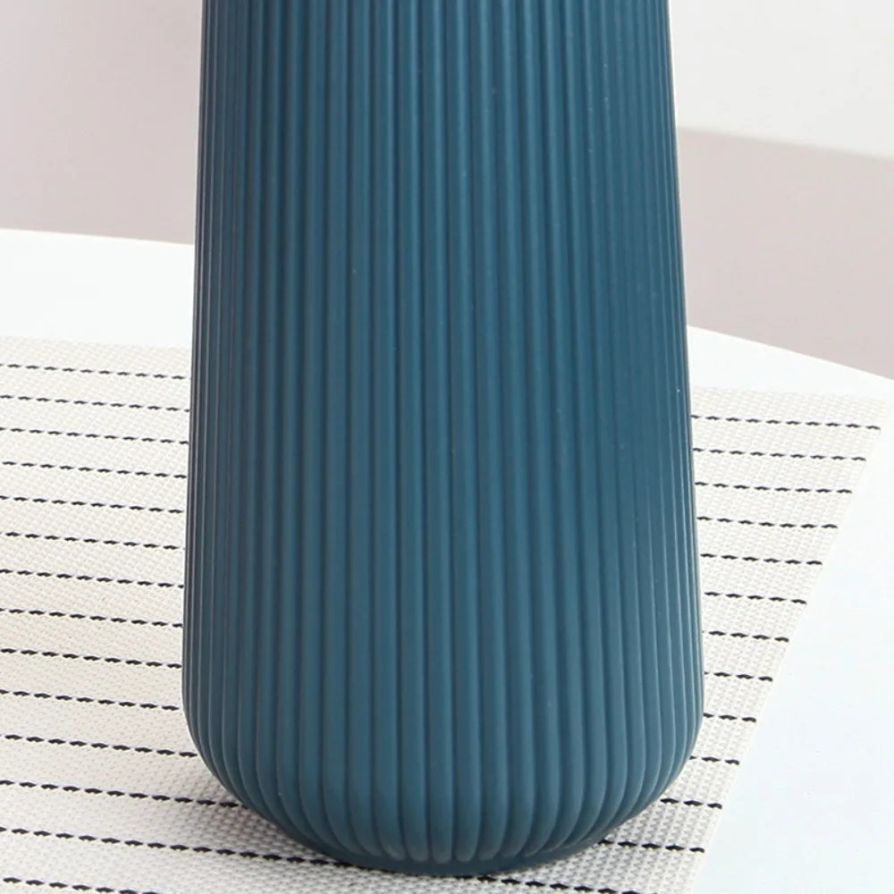 A tall, ribbed, dark teal plastic vase standing on a white surface with a black grid pattern.