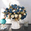 A bouquet of realistic silk blue and white roses displayed in a white vase on a dark surface.
