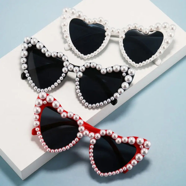 A set of pearl-embellished heart-shaped sunglasses in black and red are displayed on a white surface.