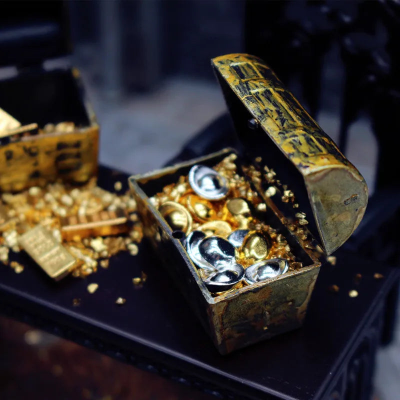 An open gold metal box filled with shiny silver and gold prosperity decorations on a dark surface.