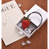 A red strawberry charm is displayed inside a white box with ice cubes and a black cord, set against a wooden background.