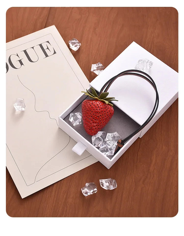A red strawberry charm is displayed inside a white box with ice cubes and a black cord, set against a wooden background.