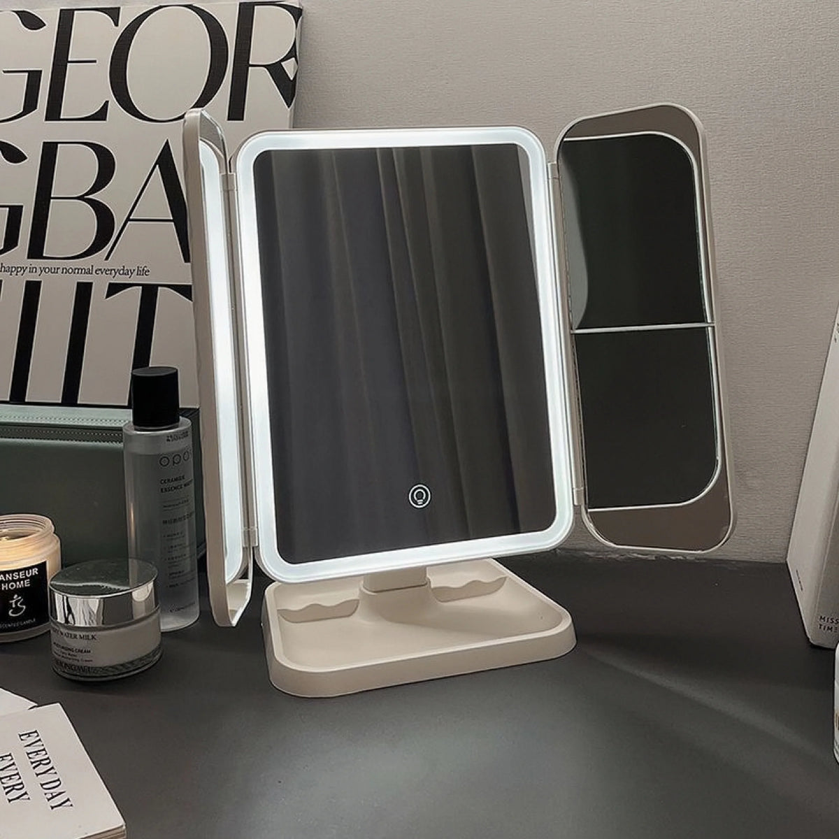 A triple fold LED makeup mirror with a light frame and a reflective surface is displayed on a dark surface.