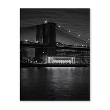 A black and white nightscape photograph of the Brooklyn Bridge spanning a body of water toward the Manhattan skyline.