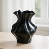 A black ceramic vase with a textured wave pattern sits on a light wooden surface near a window.