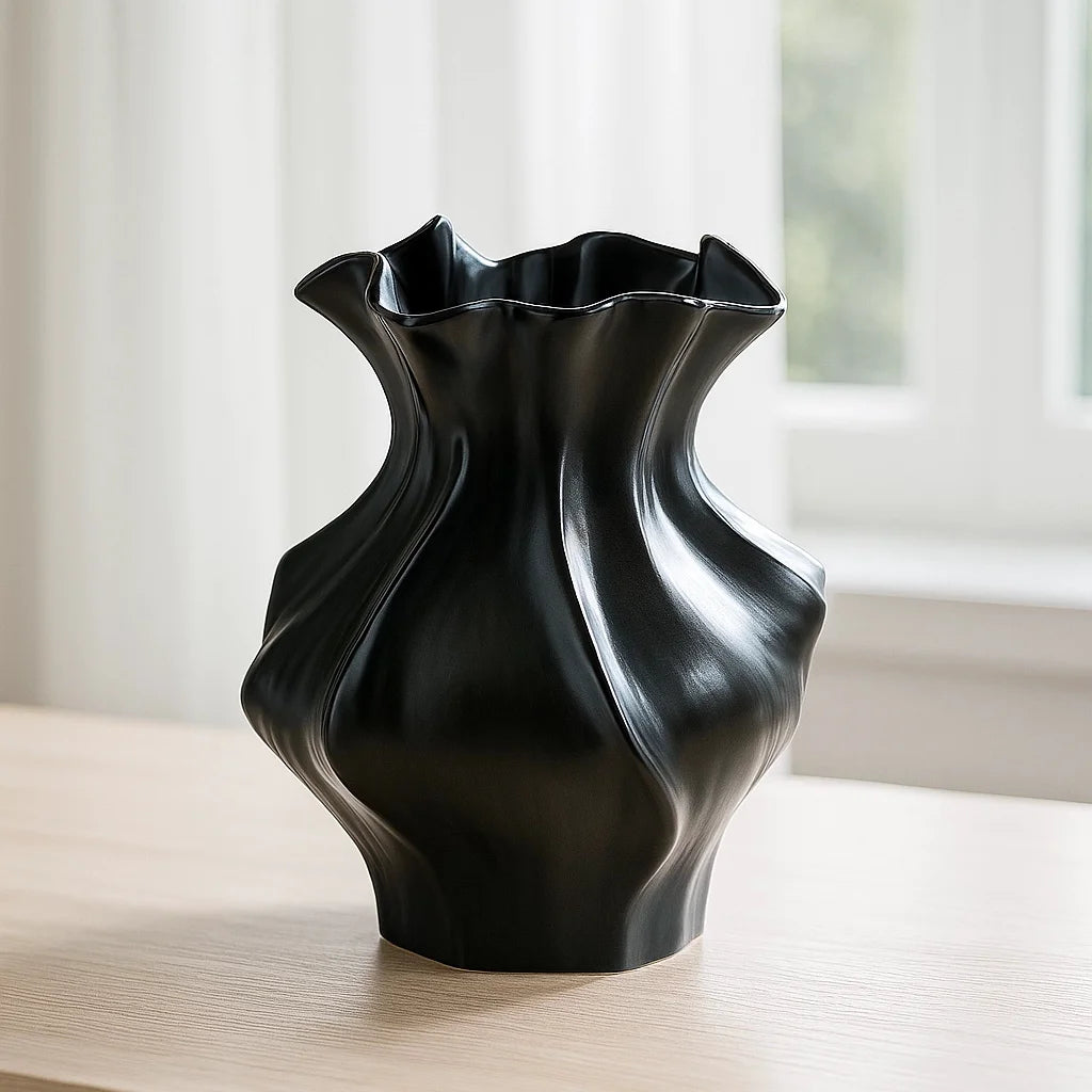 A black ceramic vase with a textured wave pattern sits on a light wooden surface near a window.