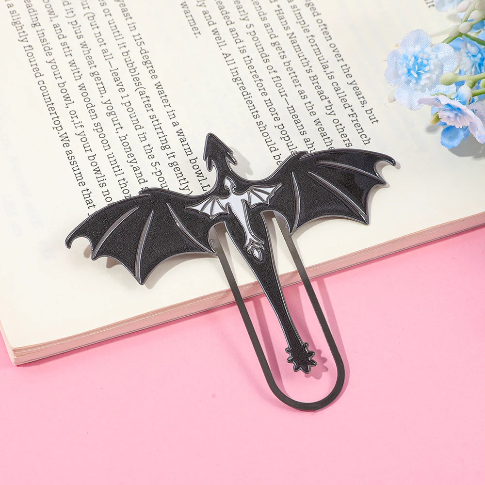 A black metal bookmark shaped like a four-winged dragon is placed on a pink surface next to an open notebook.