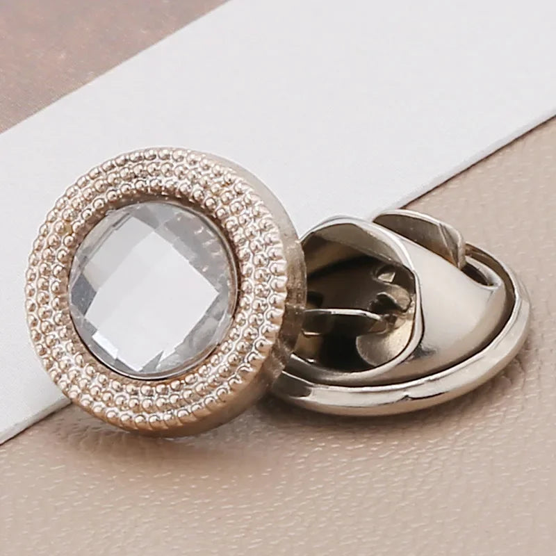 A close-up of a round, clear rhinestone button with a textured gold rim and a silver clasp.