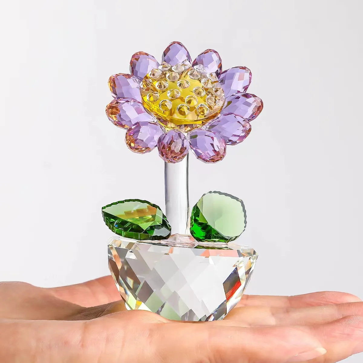 A handcrafted glass crystal sunflower figurine with a purple and yellow flower head and green leaves resting on a hand.