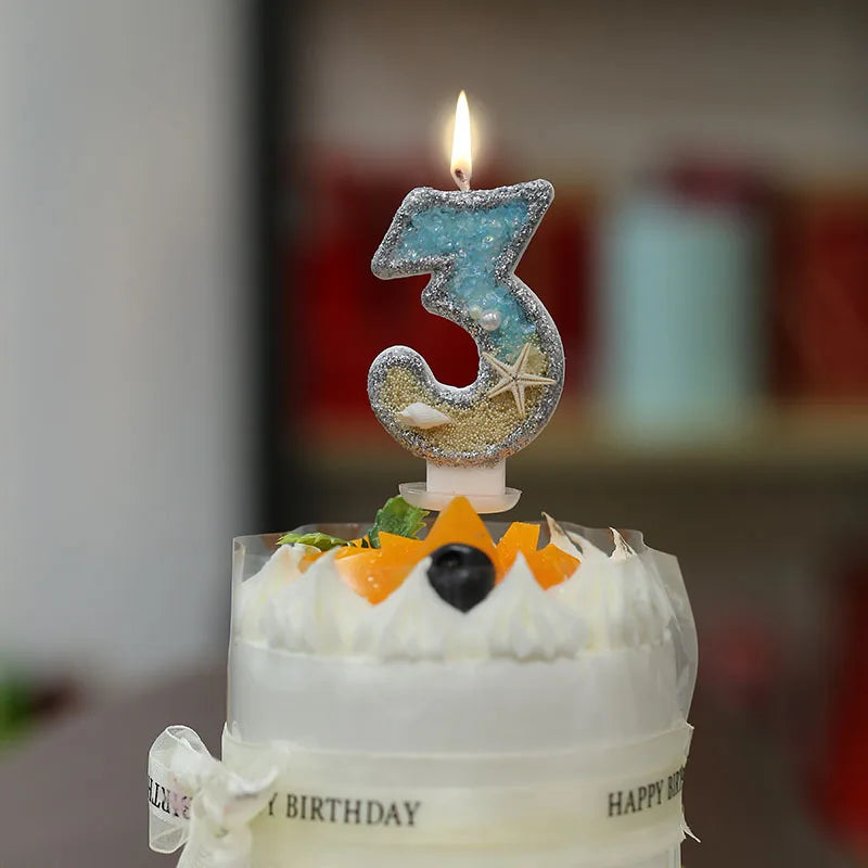 A white cake with a glitter number three topper featuring a starfish design and a lit candle.