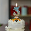 A white cake with a glitter number three topper featuring a starfish design and a lit candle.
