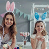 A woman and a girl smiling while wearing colorful bunny ear headbands in a light-colored room.