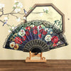 A Spanish style folding lace trim handheld fan with dark fabric and floral patterns displayed in front of a framed artwork.