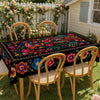 A vibrant Mexican style embroidered tablecloth set for outdoor dining with wooden chairs and a white picket fence backdrop.