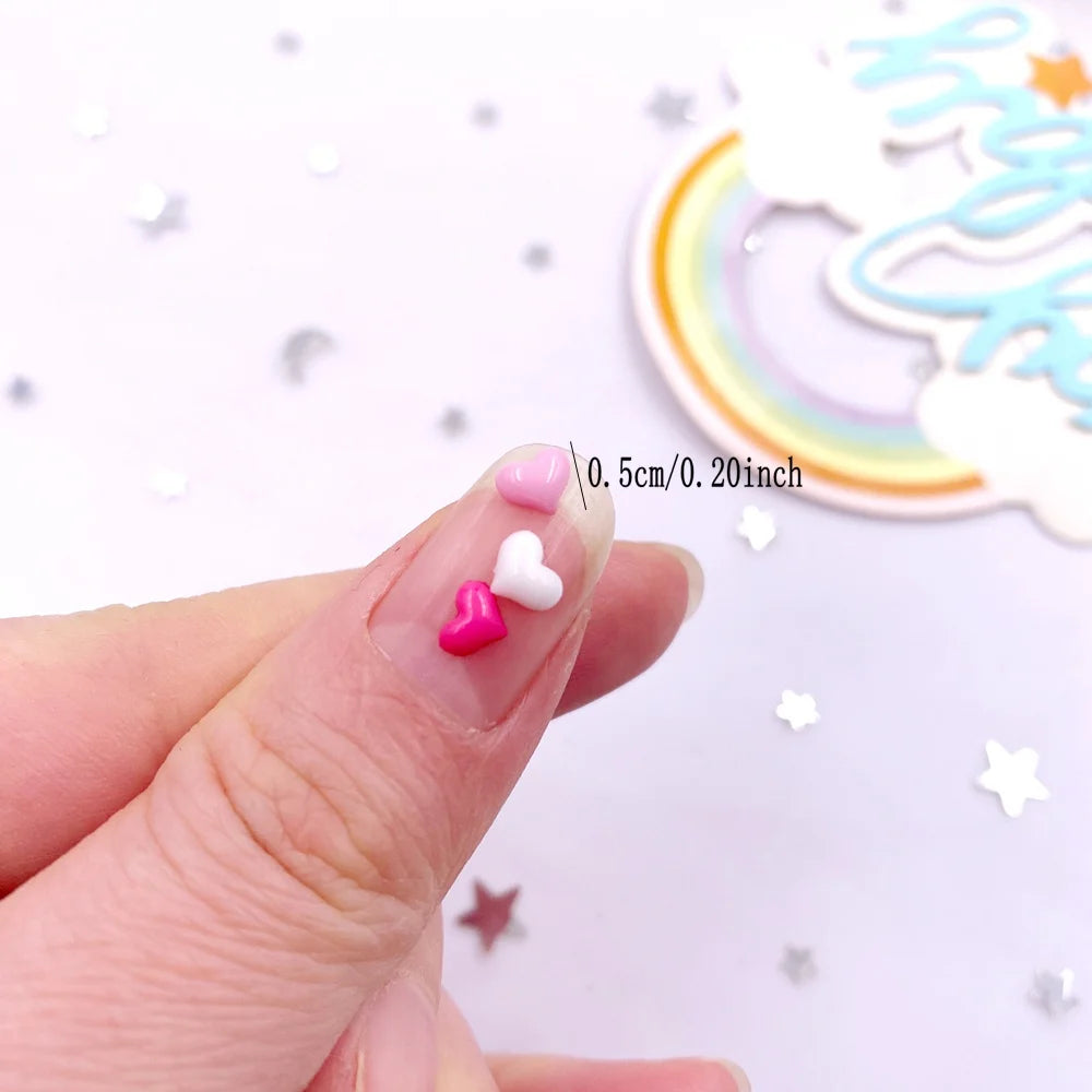 A hand holds two small pink and white heart-shaped flatback rhinestones next to a rainbow-shaped cutout on a white background
