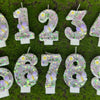 A display of number cake candles decorated with glitter, butterflies, and flowers on green grass.