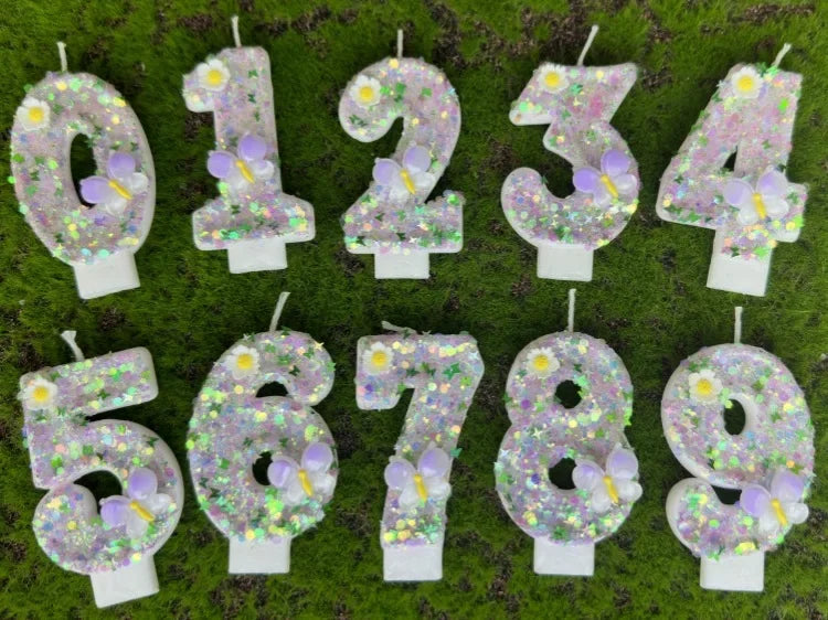 A display of number cake candles decorated with glitter, butterflies, and flowers on green grass.