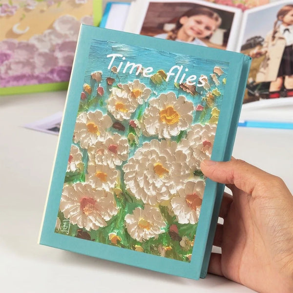 A hand holds a teal photo album titled Time Flies featuring watercolor flowers.