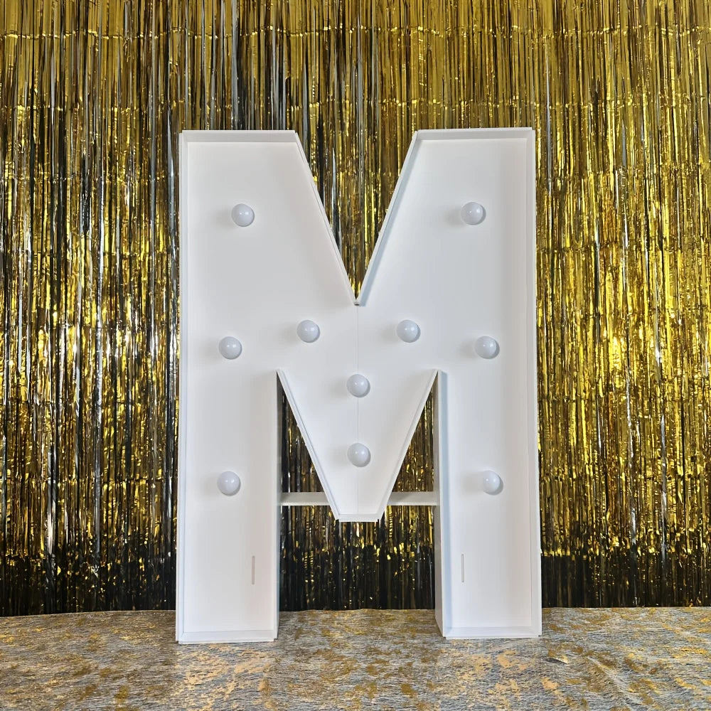 A large white marquee letter M with round lights displayed against a gold metallic curtain background.