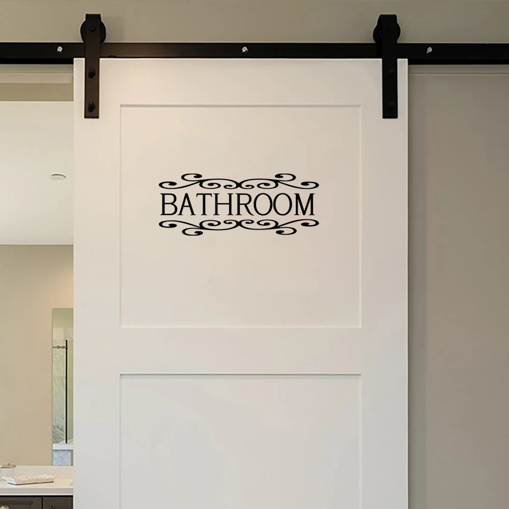 A white rectangular sign with the word BATHROOM in black script is mounted on a wall.