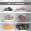 Various fresh foods like strawberries, herbs, and vegetables are stored in clear reusable cling film in white bowls.