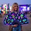 A woman holds a black folding hand fan decorated with colorful fluorescent mushrooms and glowing patterns at dusk.