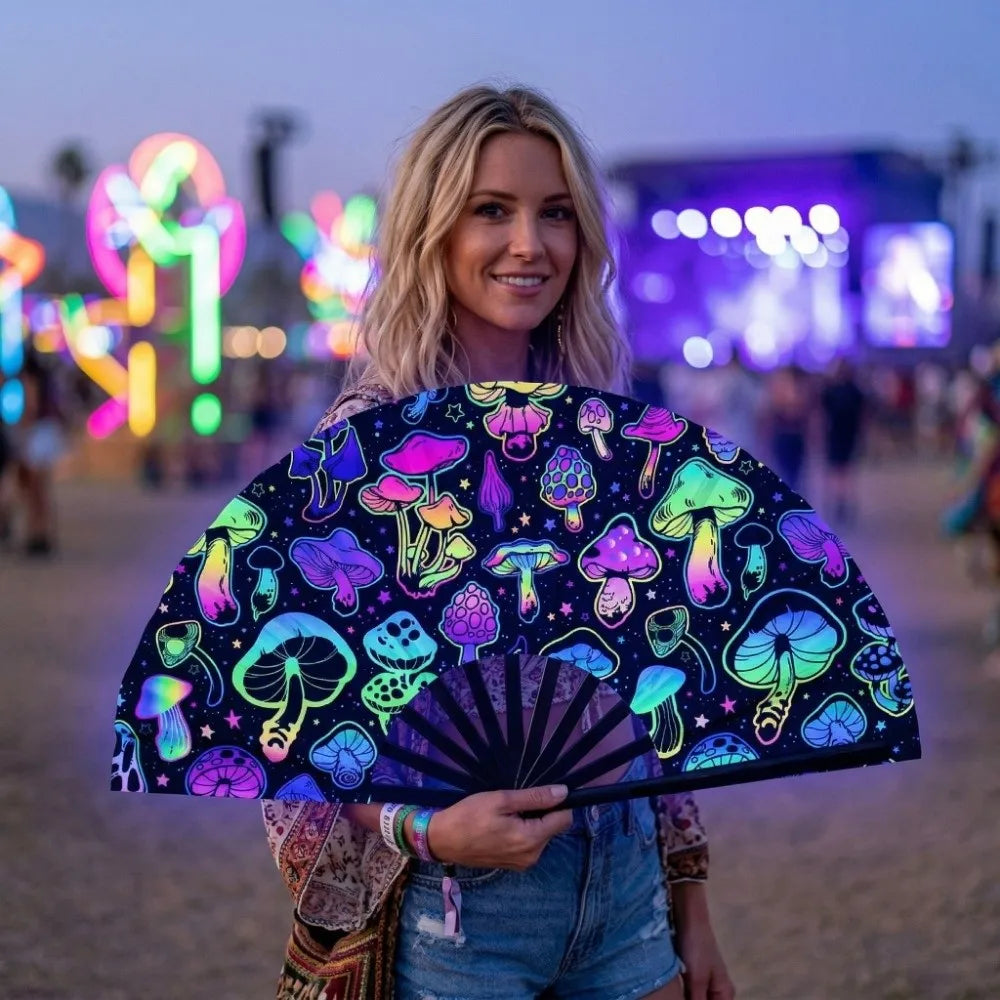 A woman holds a black folding hand fan decorated with colorful fluorescent mushrooms and glowing patterns at dusk.