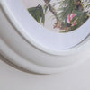 A white oval picture frame with a natural green and pink foliage pattern is shown from above.