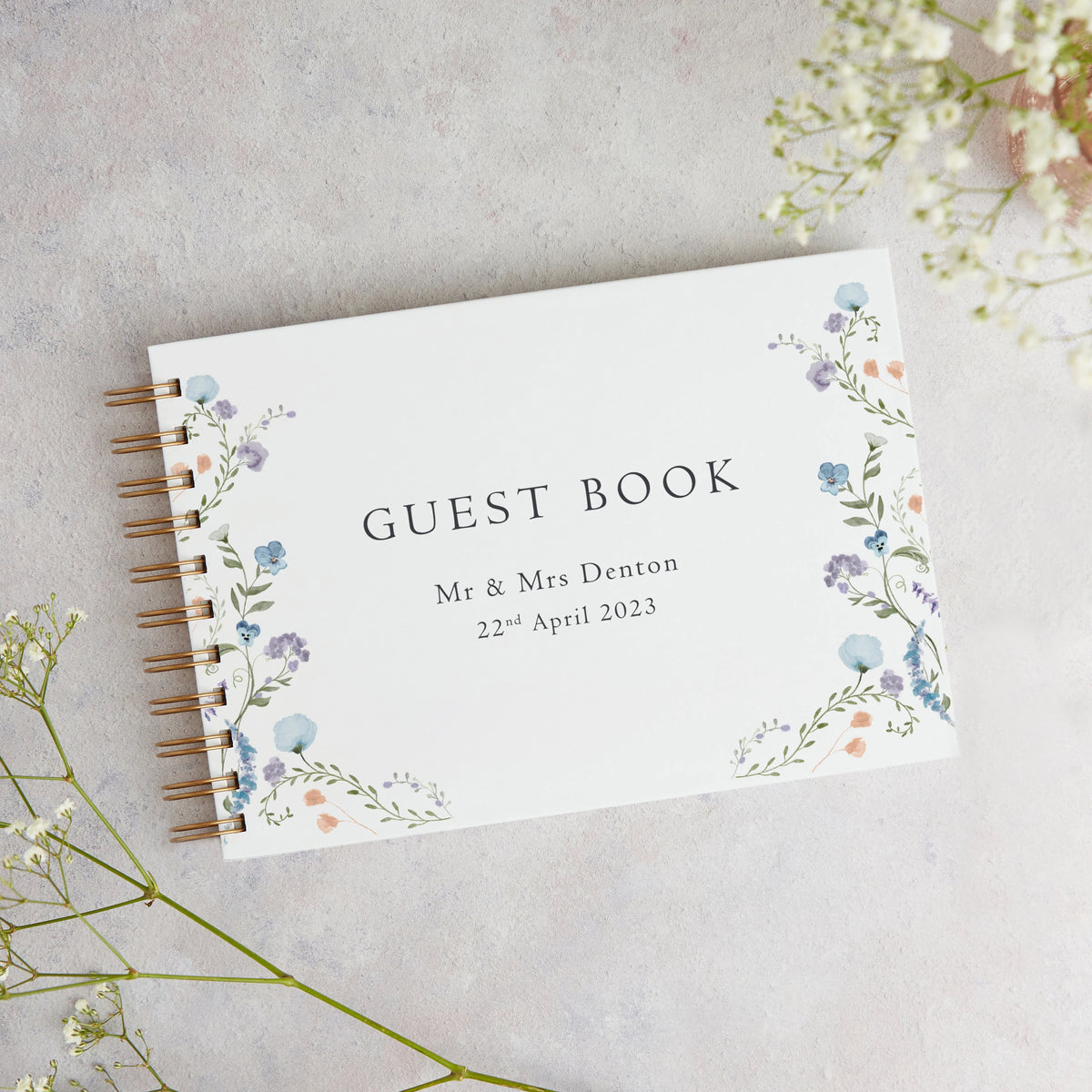 A white personalized wedding guest book with floral watercolor accents and a gold-colored spiral binding on a light gray back