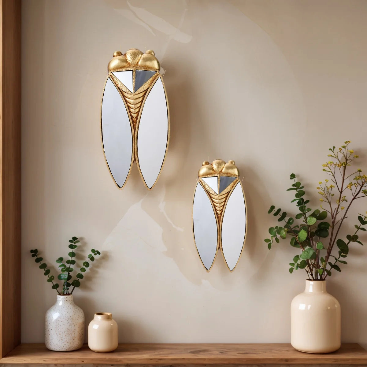 A pair of golden cicada-shaped hanging ornaments with silver accents mounted on a beige wall above a wooden shelf.