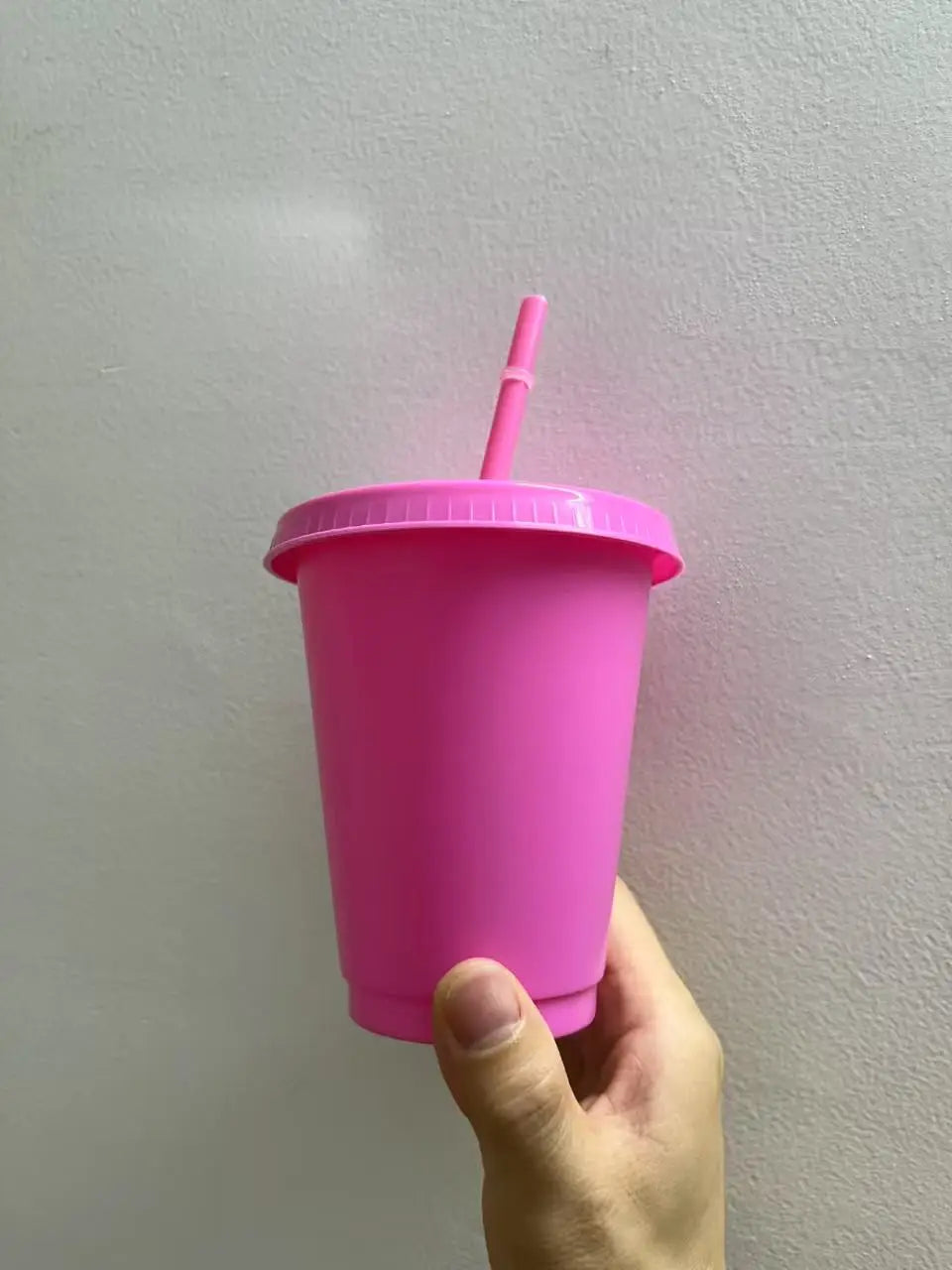 A hand holds a bright pink plastic cold cup with a pink lid and straw against a white wall.