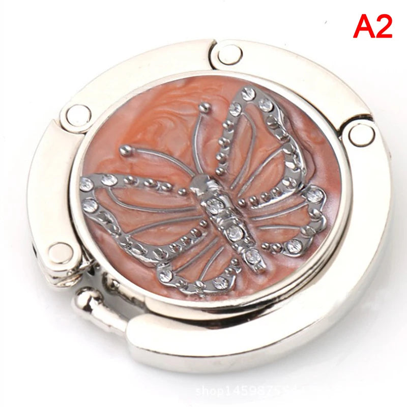 A silver foldable handbag hook with a butterfly design on a pink background.