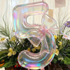 A large, iridescent, transparent number balloon shaped like the number 5 is displayed in a festive floral arrangement.