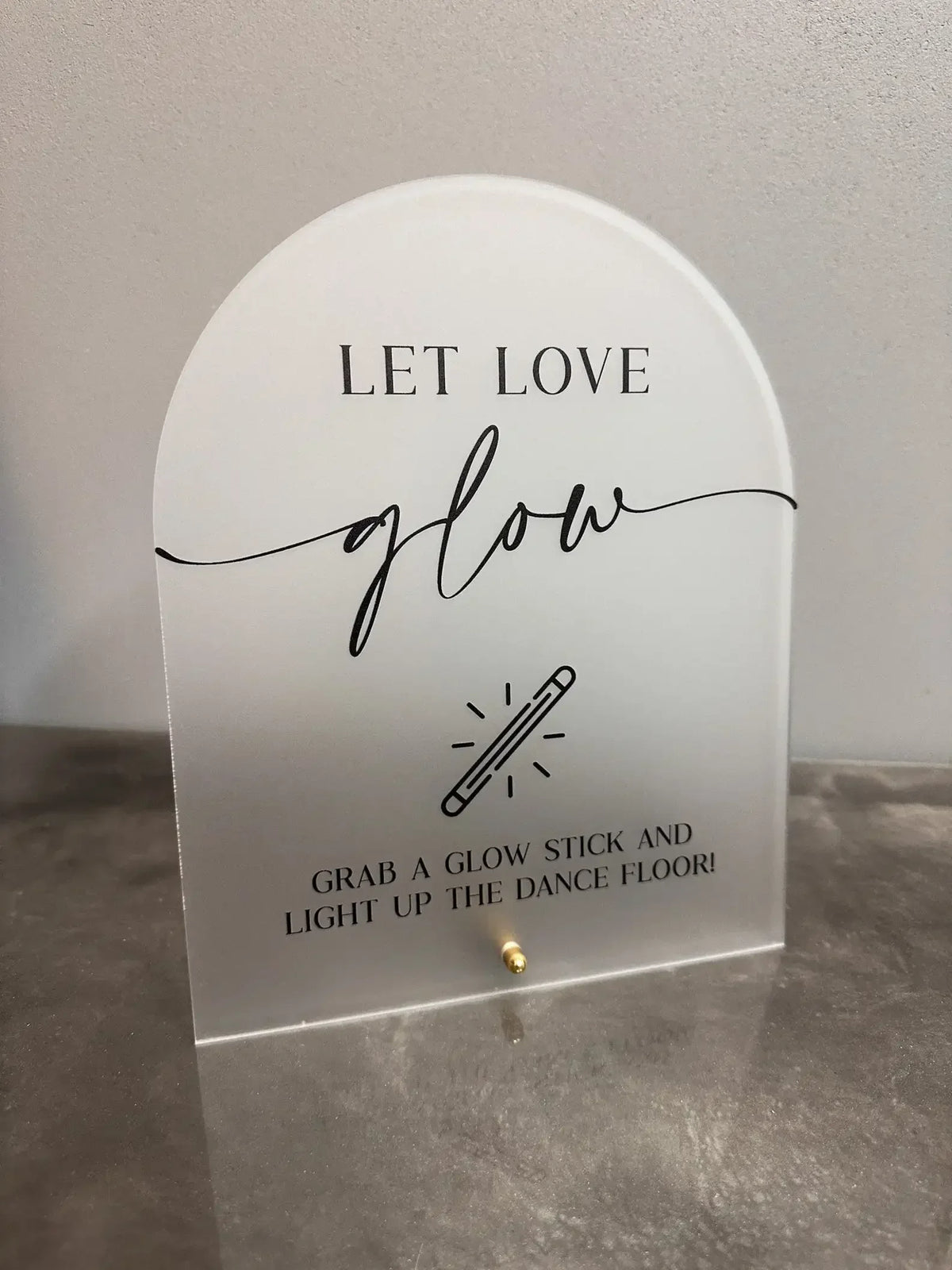 A white acrylic sign with black script that reads Let Love glow and features an illustration of a glow stick.