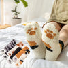 A pair of white plush socks with brown cat paw prints are displayed on a white bed with soft lighting.