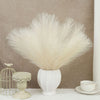 A white artificial pampas grass bouquet in a ceramic vase on a white surface with a gold wire cage nearby.