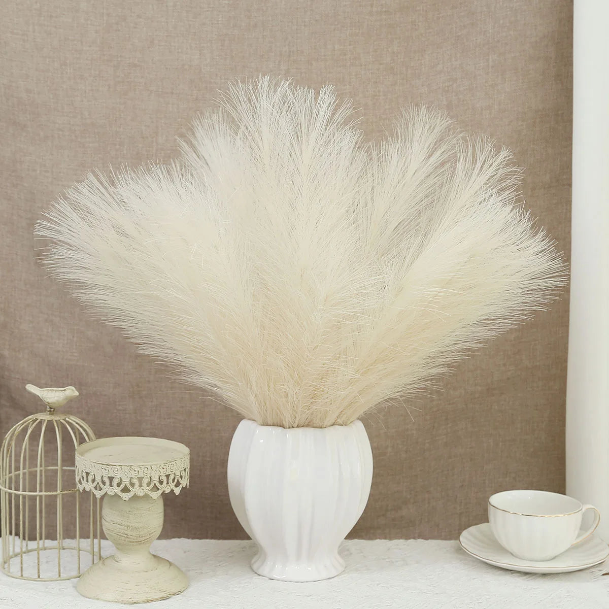 A white artificial pampas grass bouquet in a ceramic vase on a white surface with a gold wire cage nearby.
