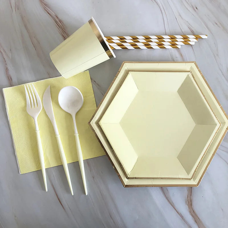 A set of gold disposable party tableware including plates, napkins, cups, and a striped straw on a marble surface.