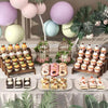 A display of various cupcakes and pastries arranged on wooden tiered stands with pastel balloons and greenery.
