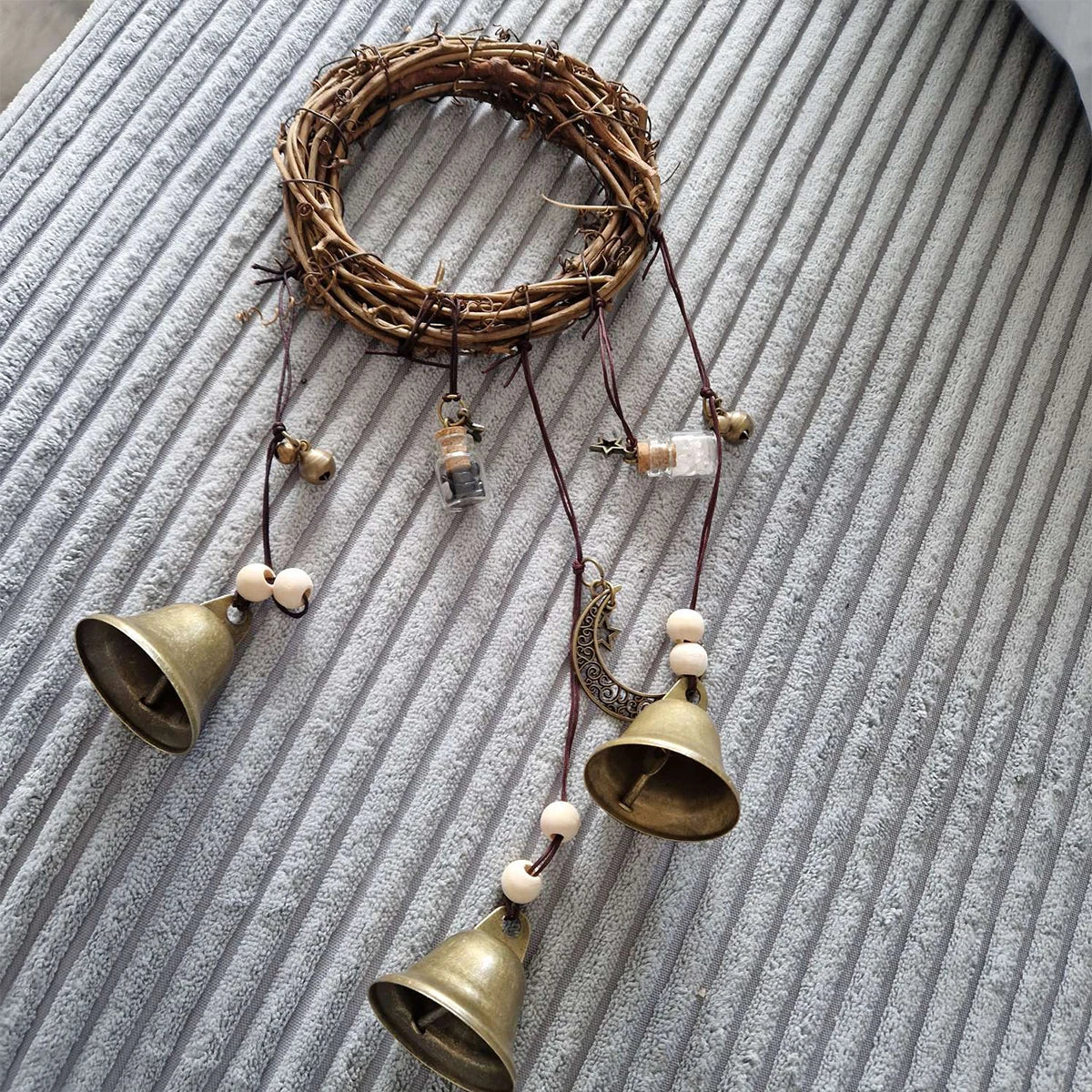 A vintage wicker wreath with hanging brass bells and beads displayed on a gray textured surface.